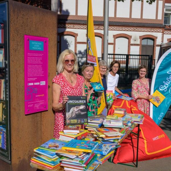 Fünf Frauen präsentieren an einem Büchertisch die gemeinsame Schön hier: Aktion, nämlich gebrauchte Kinderbücher kostenlos zu verteilen. Sie halten alle je ein Kinderbuch in der Hand. Auf dem Bild sind auch Fahnen zu sehen: Die Fahne der Lesementoren und eine Schön hier!-Fahne sowie ein roter Sitzsack von Westenergie sowie ein Teil des neuen Bücherschranks | Foto: Walter Schernstein