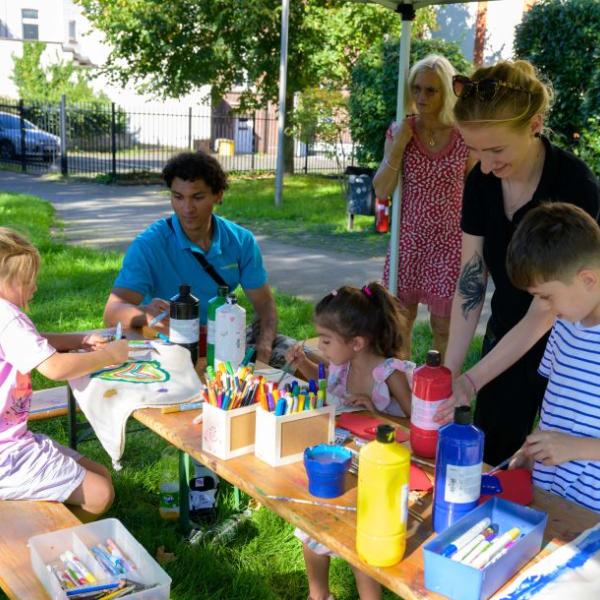 An einer Biertischgarnitur bemalen drei Kinder im Beisein von ebenfalls drei abgebildeten Erwachsenen mit bunten Farben Stofftaschen | Foto: Walter Schernstein