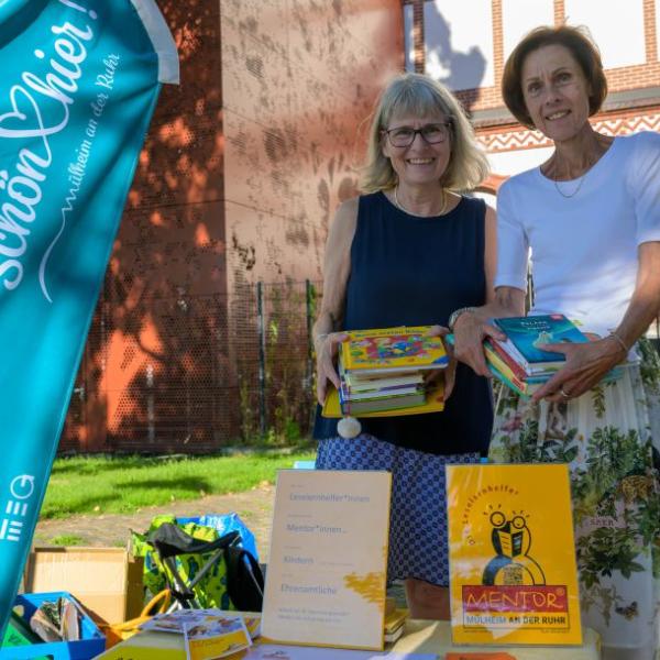 Zwei Frauen halten Kinderbücher in der Hand und stehen vor einem Tisch, auf dem weitere Bücher präsentiert werden und ein Plakat auf das Lesehelfer-Projekt Mentor hinweisen. Ebenfalls im Bild zu sehen die hellblaue Schön hier!-Fahne | Foto: Walter Schernstein