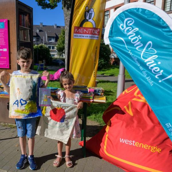 Zwei Kinder präsentieren stolz zwei selbst bemalte Stofftaschen vor einer bunten Fahnenkulisse | Foto: Walter Schernstein