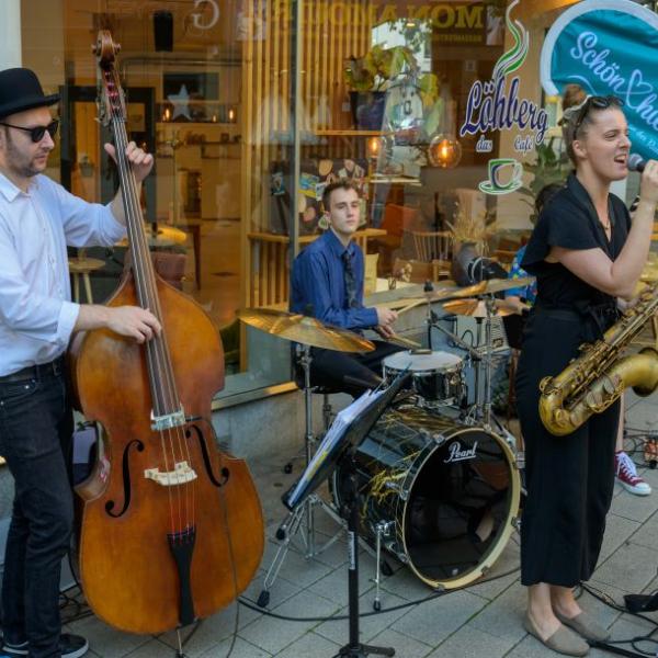 Drei Jazzmusiker*innen sind dargestellt, die vor einem Café-Schaufenster und neben einer hellblauen Schön hier!-Fahne gemeinsam auf einem Kontrabass, Saxofon und Schlagzeig musizieren | Foto: Walter Schernstein