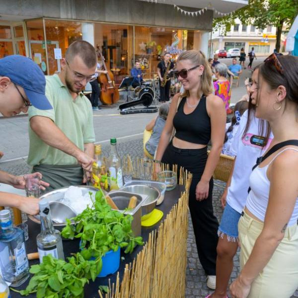 An einem Cocktailstand bereiten zwei junge Männer Cocktails für drei junge Frauen zu. Im Hintergrund sind Musiker zu sehen und die hellblaue Schön hier!-Fahne | Foto: Walter Schernstein