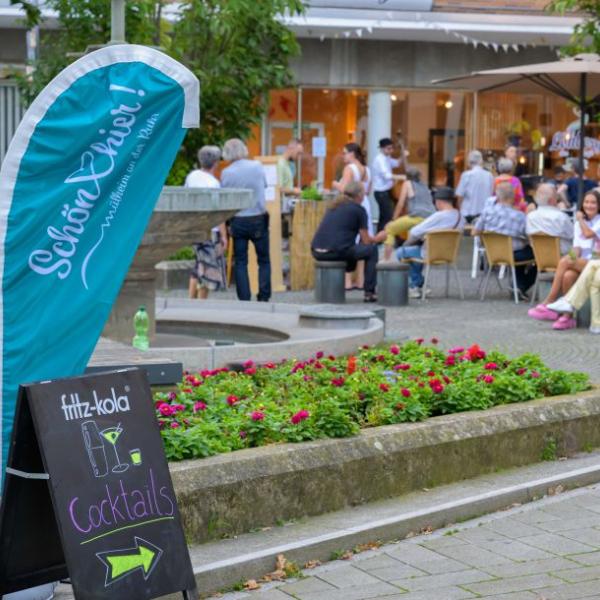 Zu sehen ist die Außenfläche des Cafés Löhberg und Teil des Brunnens Dröppelminna, an dem ein Schild aufgestellt ist, auf dem unter anderem Cocktails angepreist werden. Zu sehen ist auch eine hellblaube Schön hier!-Fahne | Foto: Walter Schernstein