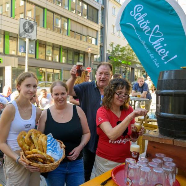 Zu sehen sind vier Personen, darunter im Hintergrund eine männliche. Zwei Personen präsentieren einen Korb mit Brezeln, eine Frau zapft Bier. Im Hintergrund ist eine hellblaue Schön hier!-Fahne zu sehen | Foto: Walter Schernstein