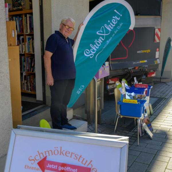 Ein Mann steht vor der Schmökerstube des Medienhauses vor der hellblauen Schön hier!-Fahne | Foto: Walter Schernstein