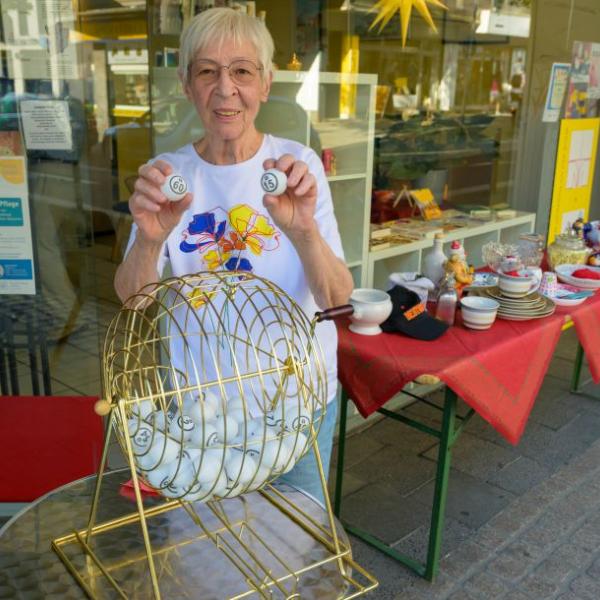 Eine ältere Dame steht vor einem Geschäft an einem Bingokorb und hält zwei Bingo-Kugeln in die Kamera | Foto: Walter Schernstein
