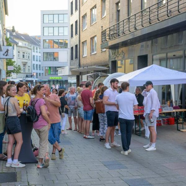 Passanten laufen eine Straße entlang, auf der ein Imbiss Waren verkauft. Andere Menschen stehen an einem Tisch gesellig beisammen | Foto: Walter Schernstein