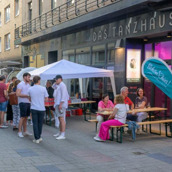 Passanten laufen eine Straße entlang, auf der ein Imbiss Waren verkauft. Andere Menschen sitzen an einem Tisch gesellig beisammen | Foto: Walter Schernstein